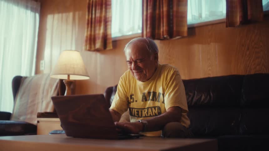 An elderly man wearing a yellow shirt sits on a dark leather sofa in a woodpaneled room typing on a laptop on a small wooden table
