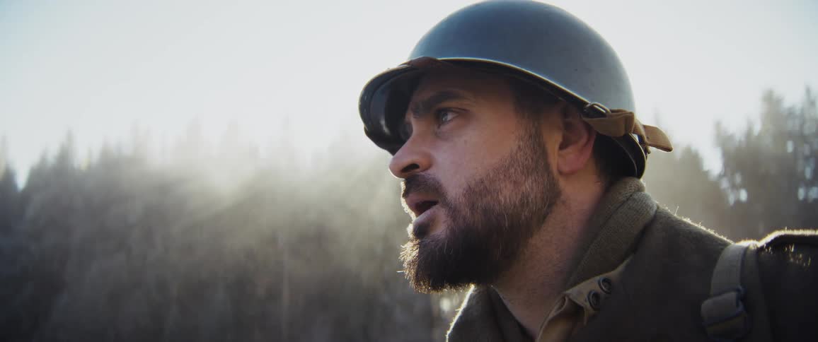 A closeup profile captures a bearded soldier wearing a vintage helmet breathing heavily in the cold air while bright sunlight creates a lens flare against a misty forest backdrop