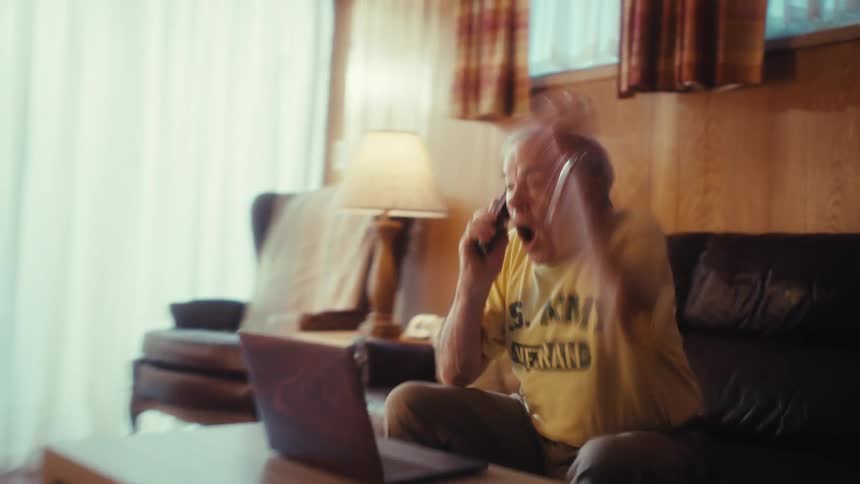 An elderly man sitting on a leather sofa in a wood paneled living room picks up a black smartphone to answer a call while working on a laptop computer