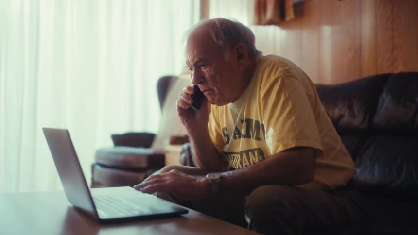 An elderly army veteran sits on a leather sofa talking on a smartphone while navigating a laptop on a coffee table sitting on a living room