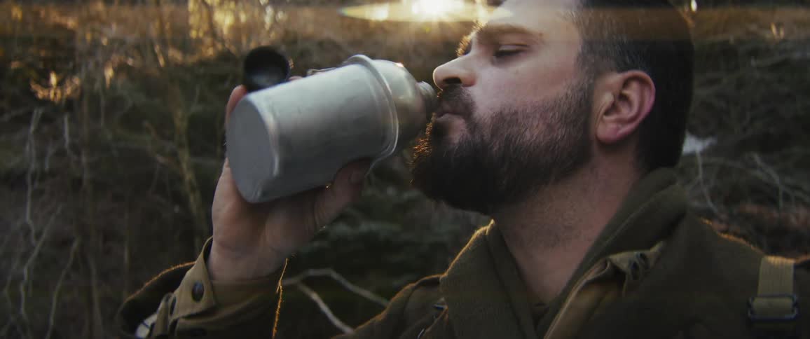 A bearded man in military attire drinks from a metal canteen in a sunlit forest then lowers the flask and looks toward the horizon while breathing heavily