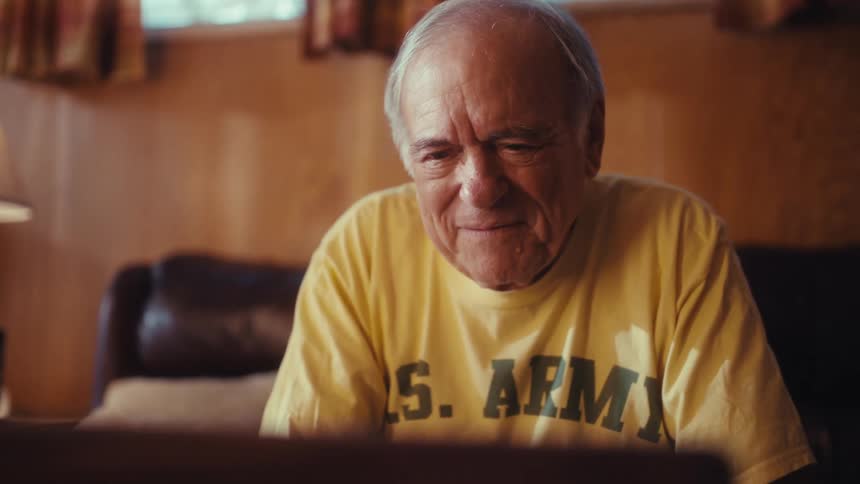 An elderly man wearing a yellow us army tshirt sits in his cozy woodpaneled living room smiling warmly and talking as he looks at a laptop screen