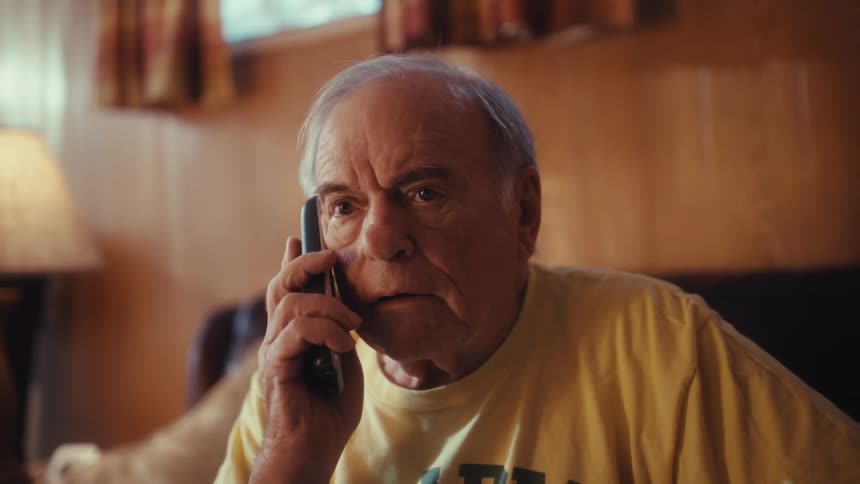 An elderly man wearing a yellow hirt sits on a black sofa picks up his smartphone to answer a call and transitions from a concerned expression to a wide joyful smile while speaking to the person on the other end