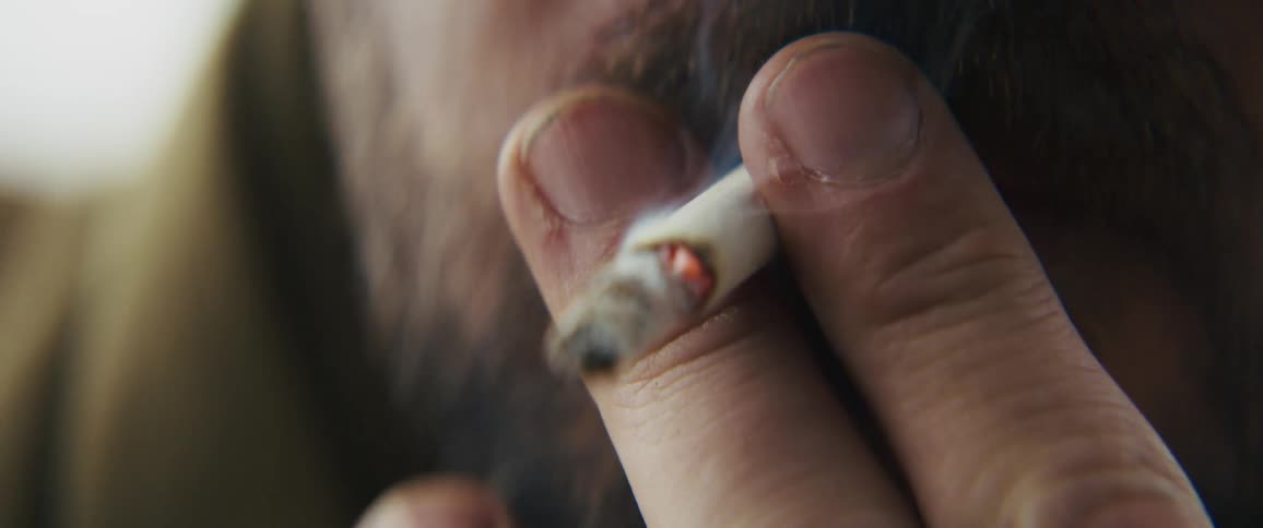 An extreme closeup shows a bearded man holding a lit cigarette to his lips while he takes a drag