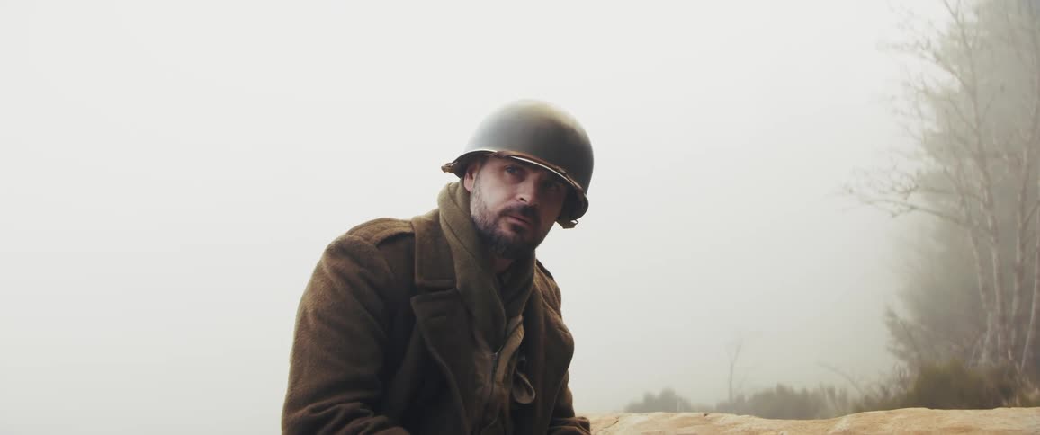 Surrounded by a thick cold mist a soldier in vintage combat gear sits on a rock looking up with a weary expression while holding a small stack of playing cards