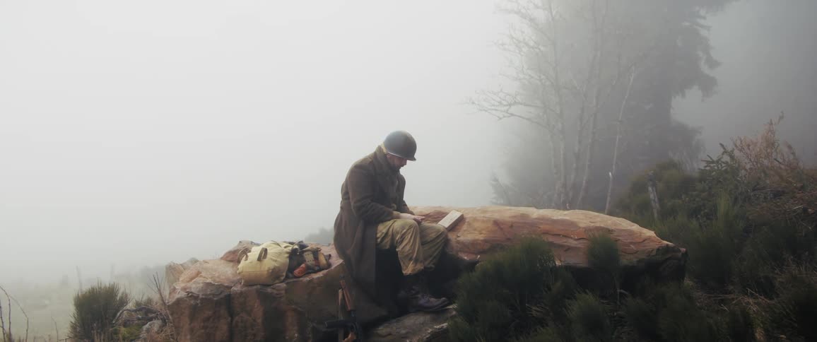 A soldier in a heavy overcoat and helmet sits on a large rock atop a foggy cliff