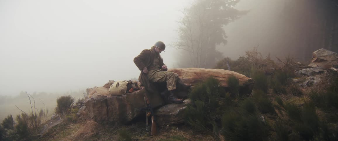 Soldier seated on a rock adjusting gear in a fogcovered mountainous landscape