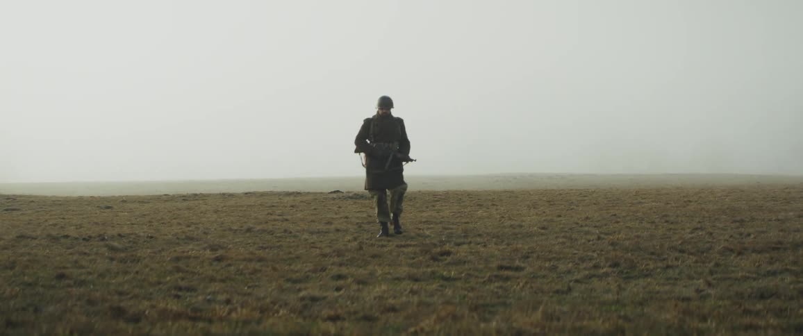 A lone soldier wearing a helmet and overcoat walks across a vast foggy field while carrying a rifle