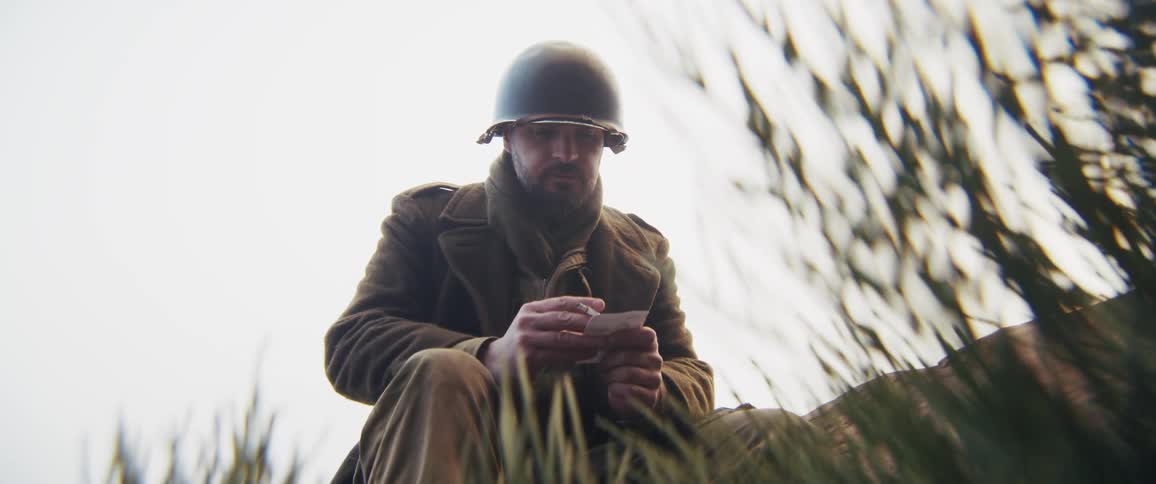 A weary soldier sits amidst tall grass smoking a cigarette while gazing at a small photograph before tucking it away and burying his face in his hands