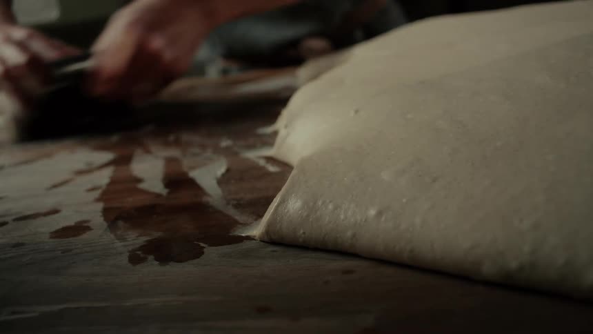Bread dough resting on a wooden surface while another portion is being handled in the background