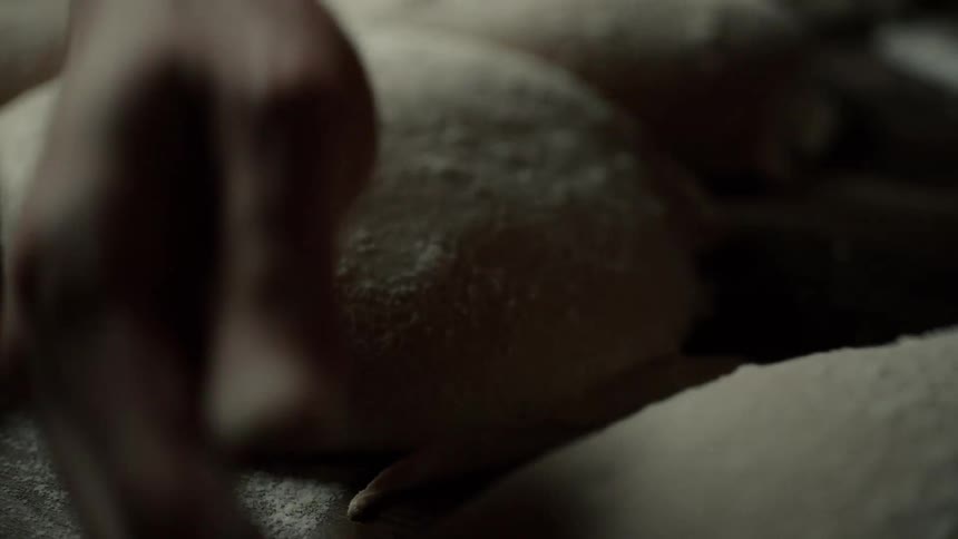 A baker picking up a piece of dough from a floured surface lined with molded dough portions ready for baking
