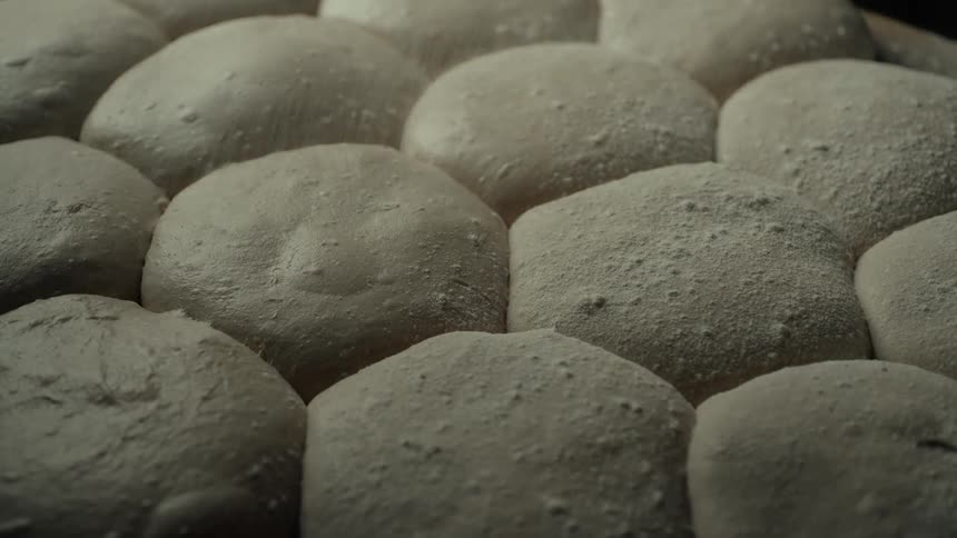 Flour being sprinkled over a series of molded dough pieces resting on a surface inside the bakery ready for the oven