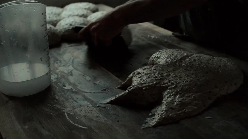 A bakers hands use a bench scraper to divide and portion seeded dough on a dark wooden workspace in a dimly lit kitchen