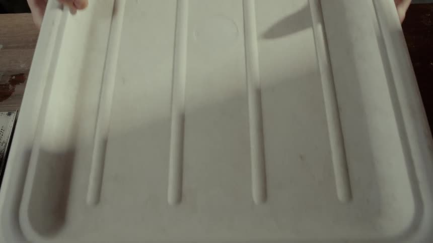 A baker places a white lid over a large smooth mound of dough resting inside a square bin before lifting the container off the flourdusted wooden workbench