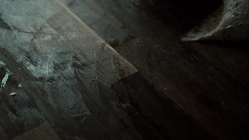 In a dimly lit kitchen a baker firmly scrapes sticky wet dough residue off a rustic wooden workbench using a metal tool clearing the surface with practiced efficiency
