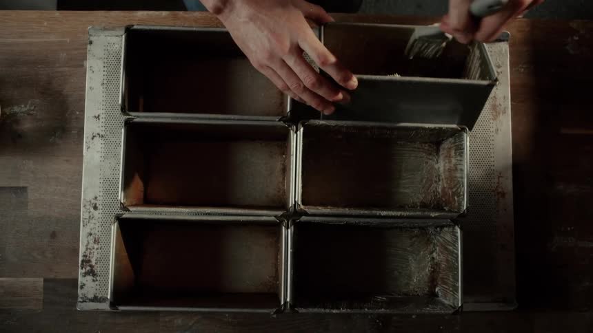 Viewed from above a baker uses a brush to thoroughly grease the inside of rectangular metal bread tins arranged in a grid on a rustic wooden table