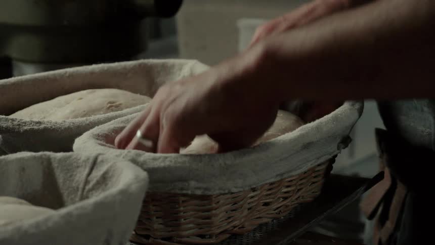A bakers flourdusted hands carefully place a shaped loaf of dough into a lined proofing basket pressing it gently into place and lifts a tray full of baskets to another place