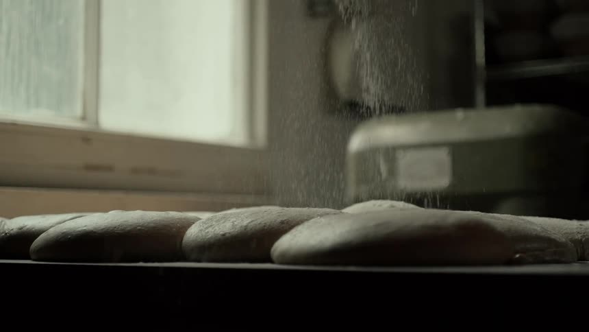 A cloud of fine white flour rains down gently onto rows of raw dough coating the smooth rising loaves in a delicate layer of dust while soft natural light spills in from a nearby window