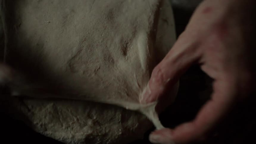 A large soft mass of dough is slammed onto a dark wooden table before a bakers hands expertly stretch fold and tuck it into a smooth round loaf