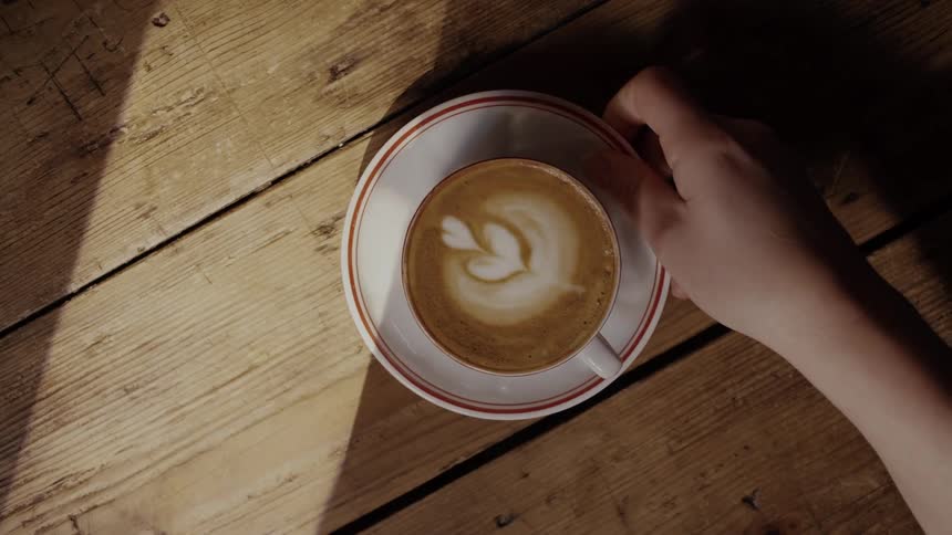 A hand gently places a fresh cappuccino featuring delicate latte art onto a rustic wooden table bathed in bright angular sunlight that casts deep shadows