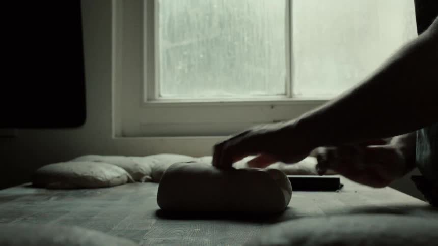 Silhouetted against the bright light of a kitchen window a baker stretches and folds a portion of soft dough using a metal scraper to skillfully shape it into a tight round loaf on the wooden workbench