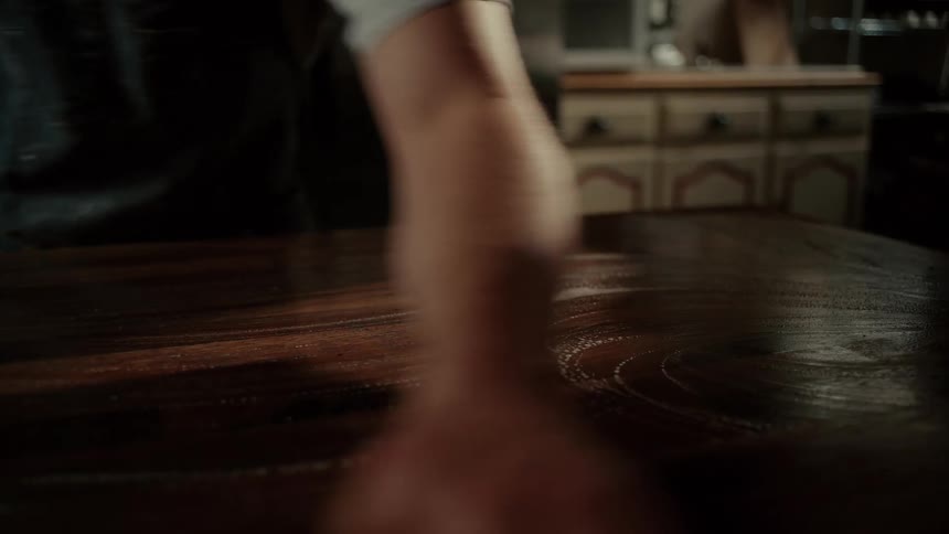 A bakers arm moves rhythmically across a rich dark wooden table wiping the surface clean with a cloth in broad sweeping motions within a dimly lit kitchen