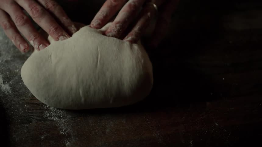 A bakers hands expertly slam a mound of soft dough onto a dark flourdusted wooden table stretching and folding it repeatedly until it forms a smooth tight loaf