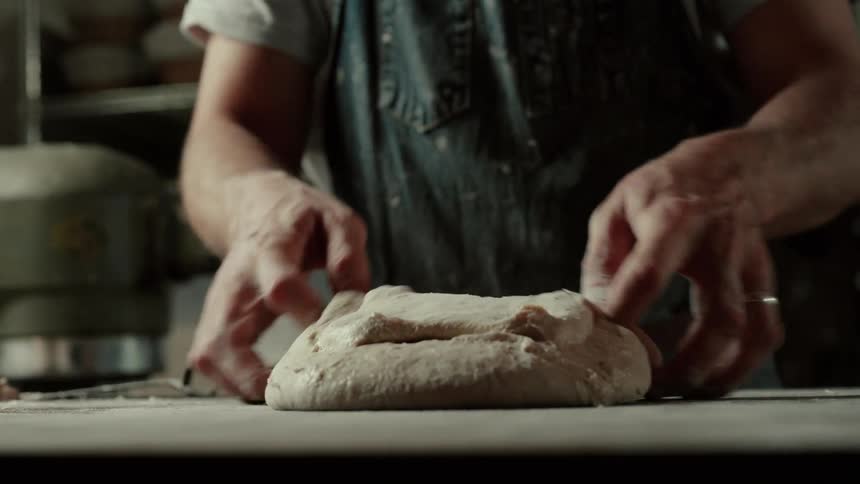 Wearing a denim apron a baker uses a metal scraper and his hands to stretch and fold soft dough on a floured workbench skillfully shaping it into a smooth round loaf in front of a heavy mixer