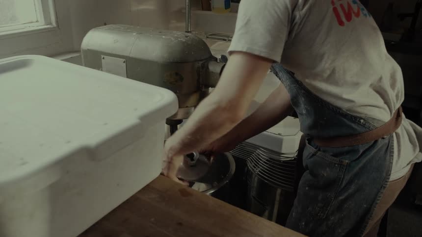 A baker leans over a large industrial machine using his hands to forcefully pull and scrape thick dough off the metal mixing blade