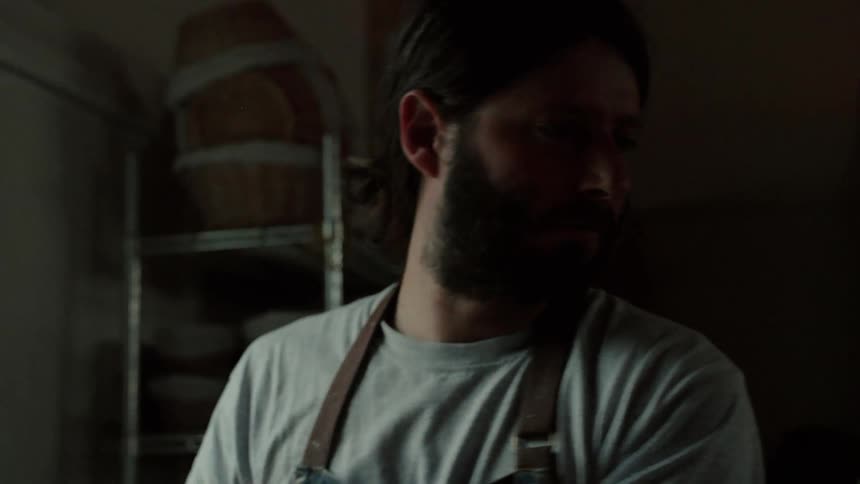 A bearded baker wearing a grey tshirt and apron works with focused intensity in a dimly lit kitchen casting his gaze downward while wire shelves hold stacked baskets behind him