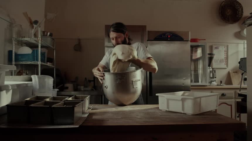 A male baker in an apron works in a kitchen reaching into a large metal mixing bowl to lift out a massive piece of pale dough and carefully placing it into a white plastic container on a wooden workbench surrounded by baking equipment