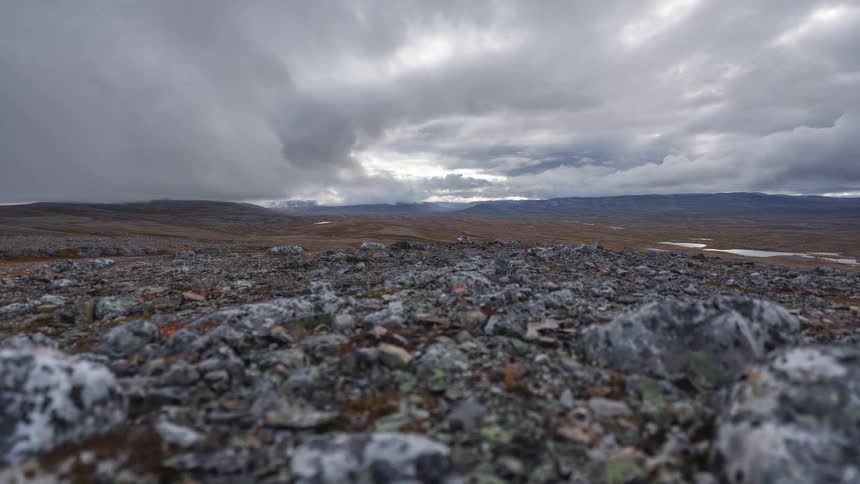 Dark clouds move over a vast and desolate rocky plateau while sunlight breaks through the horizon in the distance