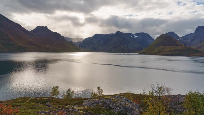 Mountains on the lake as plants and rocks overlook the water under a cloudy sky