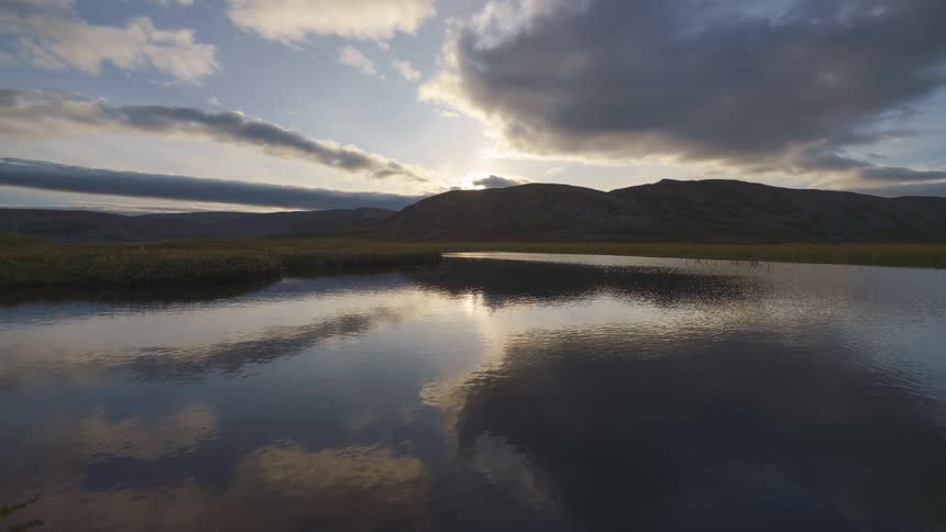 A lake view with a mountain rising from the shoreline the sun setting beneath its peak under a cloudy sky