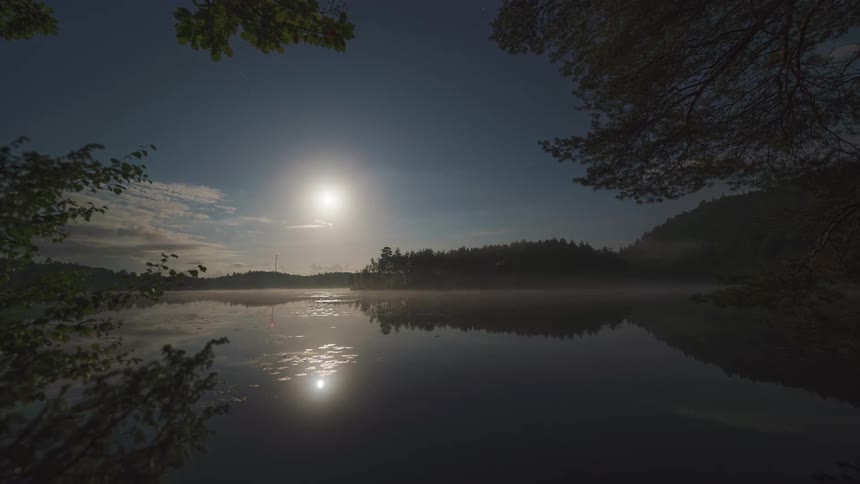 A still lake surrounded by trees with an orange setting sun glowing through a cloudy evening sky
