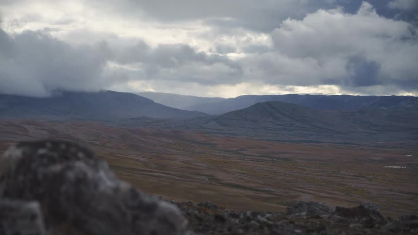 Thick clouds move over a vast and desolate tundra valley bordered by dark rugged mountain ranges under a heavy gray sky