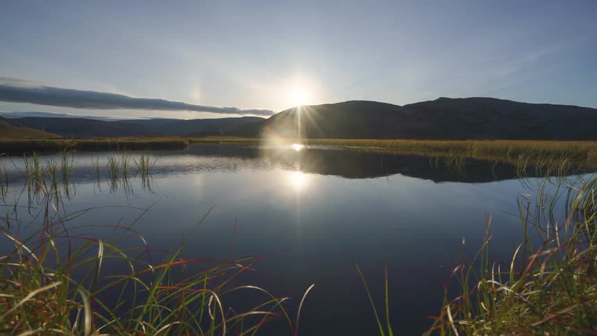 A bright sunburst dips behind a dark mountain ridge casting a brilliant reflection across the calm lake while the camera glides through tall green marsh grasses