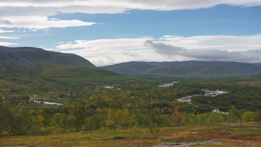 A wide shot captures a winding river flowing through a lush valley of autumn colored trees bordered by large rolling mountains under a bright sky with scattered clouds in a quiet remote wilderness area during the daytime