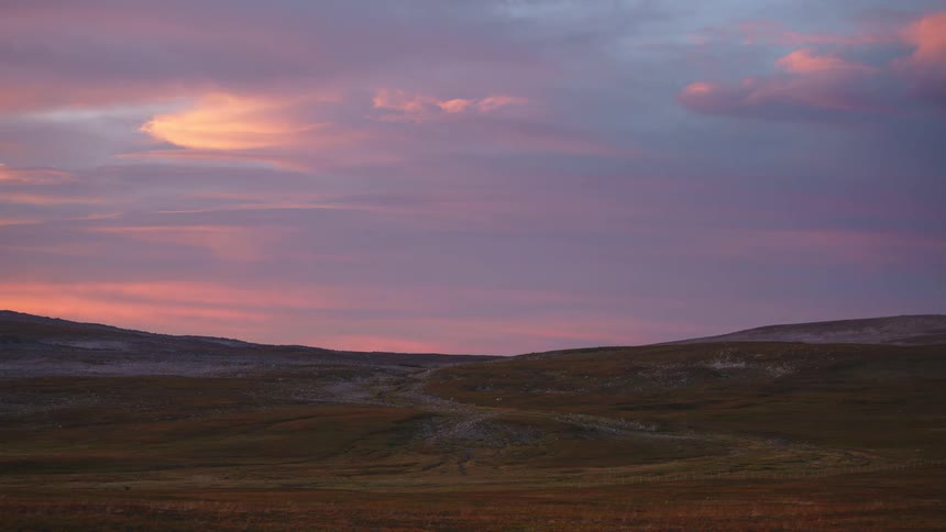 Soft purple and orange clouds sweep rapidly across a vast twilight sky casting a fading glow over the desolate rolling hills in this serene timelapse