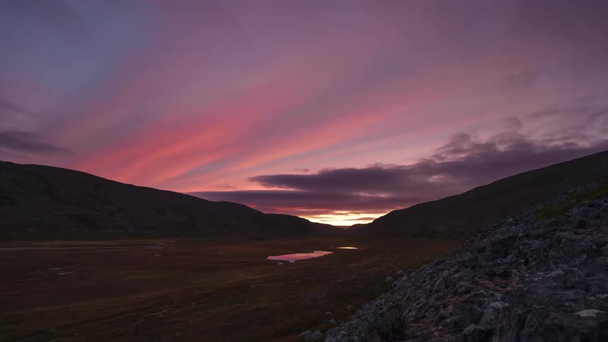 A wide timelapse captures a purple and orange sunset over a remote mountain valley where clouds drift across the sky reflecting in a small pool of water until a faint green aurora borealis begins to appear above the silhouetted peaks