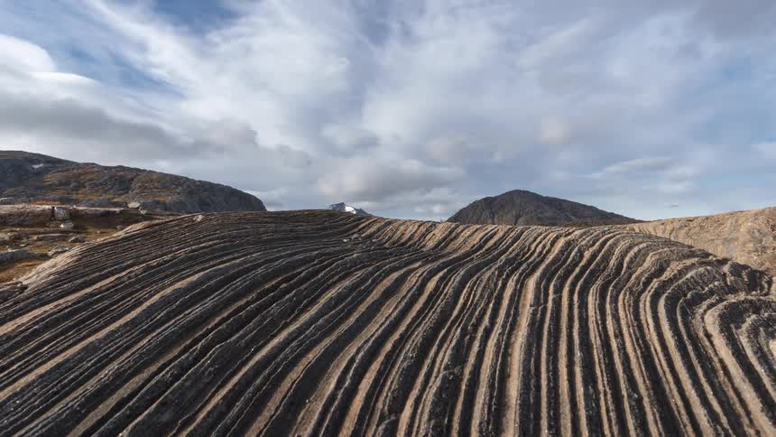 A wide shot reveals a large dark rock formation with deep parallel grooves in the foreground leading the eye toward mountains under a bright cloudy sky