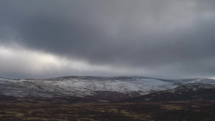 A dramatic timelapse shows heavy grey clouds rolling rapidly over snowy mountain peaks casting shifting shadows across the rugged brown tundra landscape below