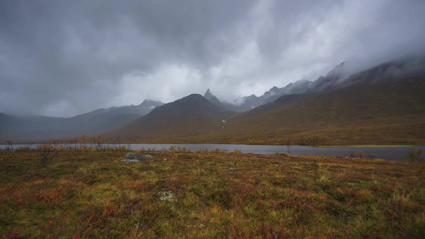 A moody timelapse captures heavy grey clouds and mist rolling rapidly over rugged mountain peaks that loom above a calm lake surrounded by autumn tundra vegetation