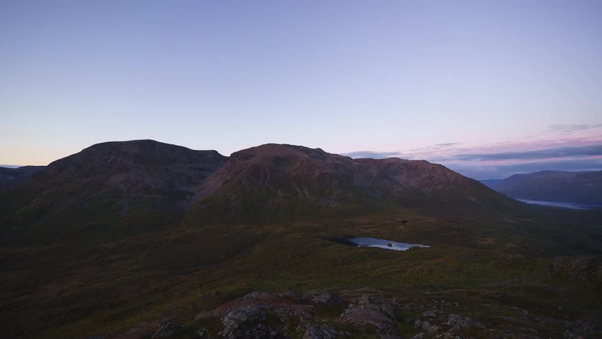 Shadows stretch across a wide mountain valley as the sun sets over a rugged tundra landscape gradually plunging the hills and a small lake into darkness while the sky shifts from blue to deep purple during the transition to twilight