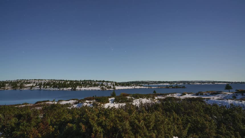 A timelapse captures the dramatic cycle of day and night over a snowy lake where the sun dips below the horizon to reveal a starry sky before rising again to bathe the landscape in bright morning light