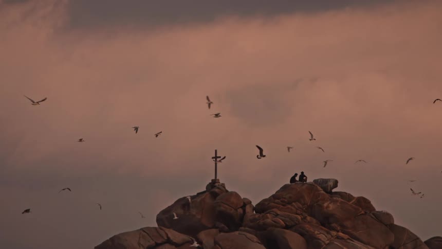 A flock of birds circles a solitary cross atop a rugged rock formation their silhouettes flitting past two figures seated against a moody cloudstreaked sky