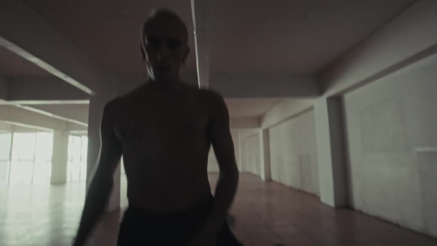 A shirtless performer walks confidently through a vast empty industrial space breaking into fluid expressive dance movements as he nears the camera