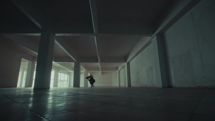 In the center of a cavernous empty warehouse a distant performer rises from the floor to execute a dramatic spin their dark flowing attire swirling amidst the stark industrial architecture