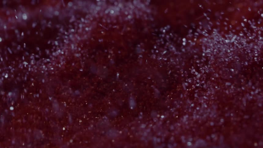 A closeup of a deep red textured surface reveals countless glittering particles nestled in soft ridges and valleys that are moving