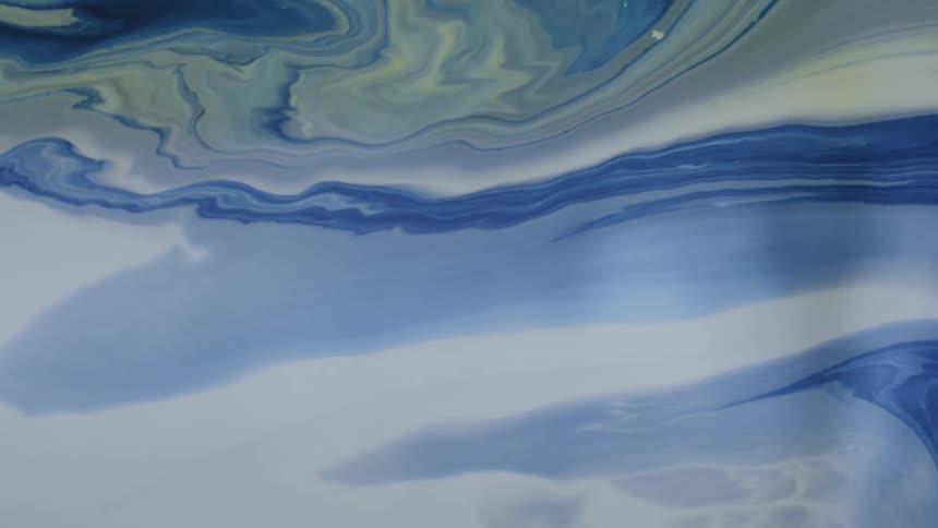 Blue and white paints swirl together in a shallow tray creating organic marble patterns and fluid ripples that slowly shift across the frame as the different liquid densities merge into abstract waves and serpentine shapes